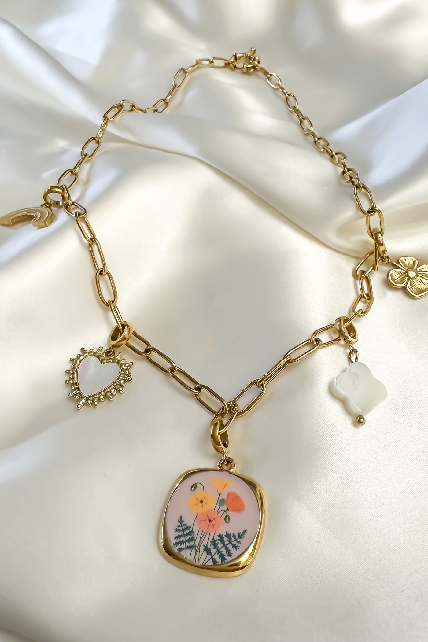 Gold paperclip chain worn as a charm necklace with a mother of pearl heart, a white clover, a gold flower, a gold rainbow and a square floral pendant, laid on cream satin