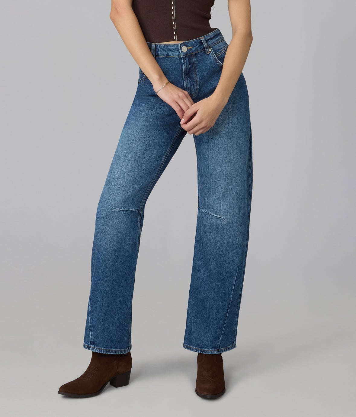 Model wearing Lola Denim LENNOX high-rise barrel jeans in a mid-blue wash with curved seams at the knee and brown suede ankle boots