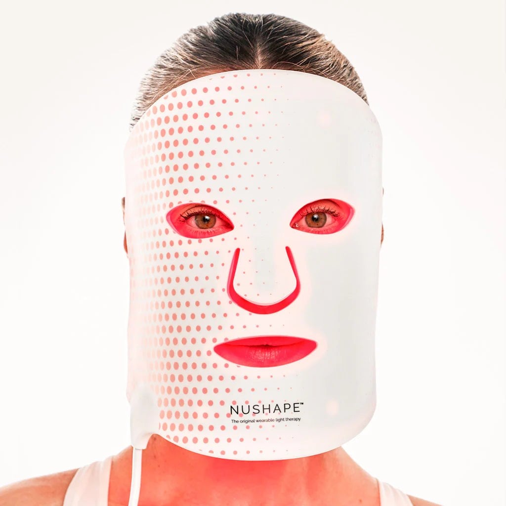 Woman wearing the white flexible silicone Nushape Red Light Therapy Mask, with red LEDs glowing through cut-outs around the eyes, nose and mouth