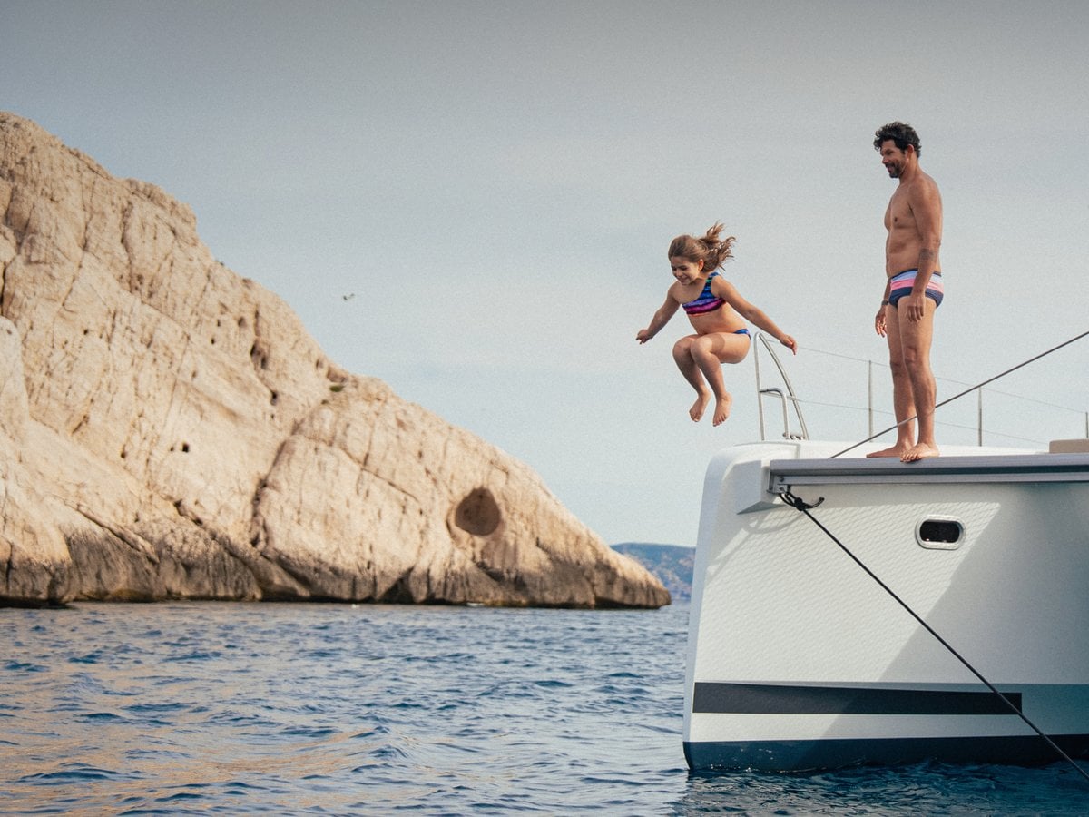 Child leaping from the bow of a white catamaran into clear water beside limestone cliffs as an adult watches from the deck