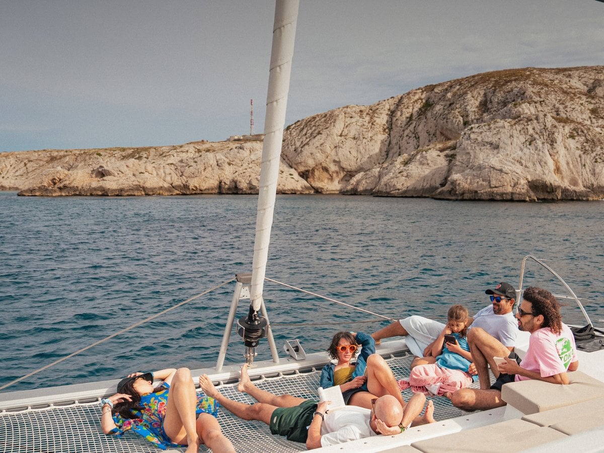 Group of friends relaxing on the trampoline net of a catamaran at anchor below rocky cliffs