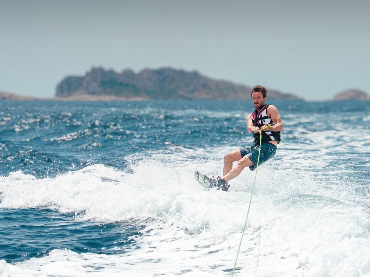 Person wakeboarding on choppy blue water behind a boat, with a rocky island on the horizon