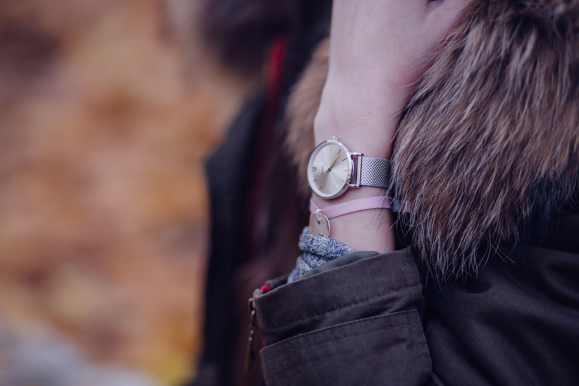 Minimalist watch with mesh band and pink bracelet worn on woman's wrist next to brown fur collar